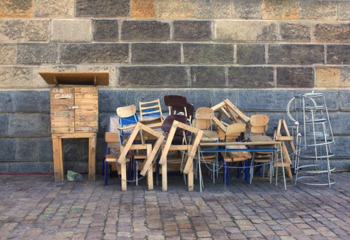 A collection of discarded chairs and small tables arranged on a cobbled pavement in front of a grey brick wall. The furniture includes wooden chairs with different styles and finishes, some with light natural wood tones, others darker or painted, and various small tables, some with thick legs and tabletops, others with thinner, metallic frames. To the left, there is a wooden cabinet or storage box with a slightly open lid, positioned against the wall, and to the right, a tall, metallic shelving unit or stand with a chrome finish. The scene suggests a collection of unwanted or scrap furniture in an outdoor area, possibly awaiting removal as part of rubbish clearance by Commercial Waste Stevenage. The environment is urban, with textured cobblestone ground and a plain, practical wall background, typical of a commercial or residential exterior in the Stevenage area.