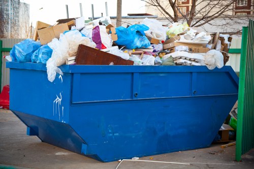 A large blue industrial skip filled with a mixed pile of waste materials, including cardboard boxes, crumpled white and purple plastics, and various paper and packaging debris. The skip is situated outdoors on a paved surface, with some green fencing partially visible on the right side of the frame. The waste appears to be loosely packed and extends beyond the top edges of the skip, indicating overfilling. The background shows a blurred urban environment with brick buildings and trees, suggesting a commercial or industrial area in or near Stevenage. This scene relates to rubbish removal services provided by Commercial Waste Stevenage, offering waste collection for businesses and local properties in the area, consistent with their service category of rubbish disposal.