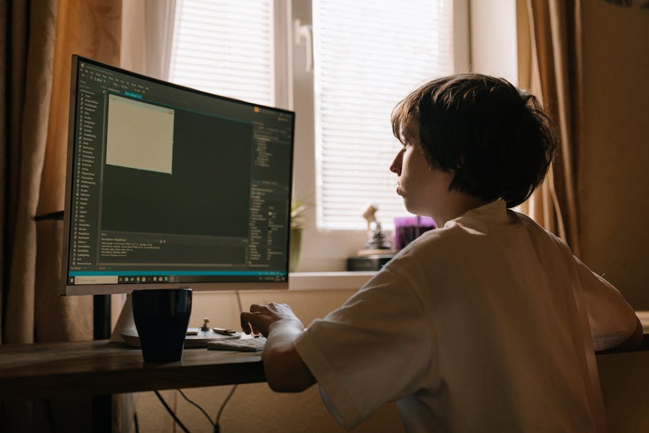A person with short dark hair, wearing a white shirt, is seated at a wooden desk in a room with natural light coming through a window with horizontal blinds. They are facing a large computer monitor d