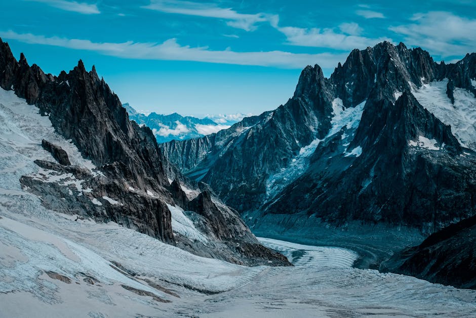 A panoramic view of a snow-covered mountain landscape featuring rugged, dark grey rocky peaks with sharp inclines and steep cliffs, some of which are dusted with patches of snow. In the foreground, a 